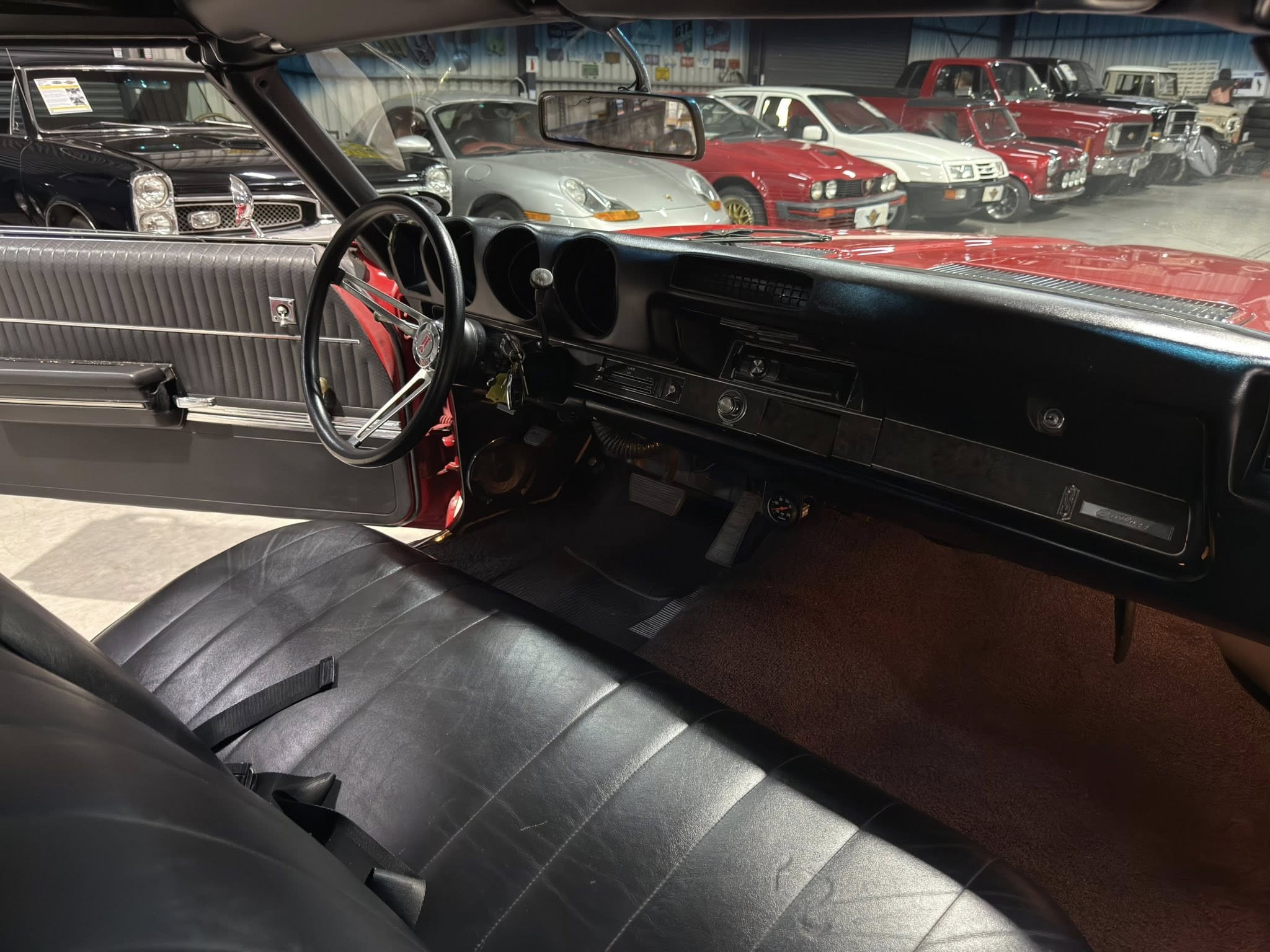 Interior of a vintage car showing the black dashboard, steering wheel, and bench seats with a showroom of classic cars visible through the windshield on display in the background?