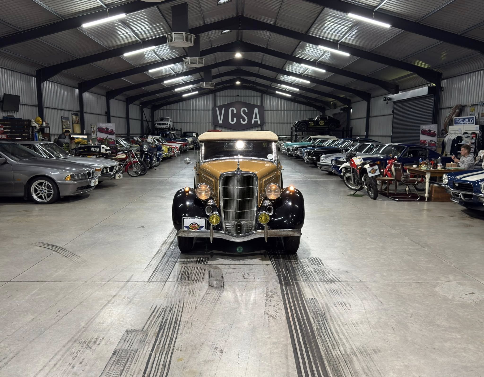 Vintage car showroom inside a large metal building; a brown classic car faces the camera, flanked by rows of vintage cars on both sides with a 'VCSA' sign in the back.