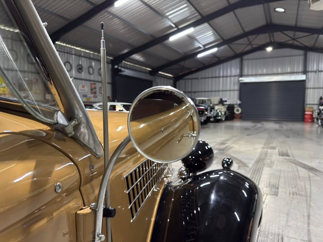 Close-up of a tan vintage car's front left fender with a round side mirror, inside a well-lit automotive showroom or warehouse with other classic cars in the background.