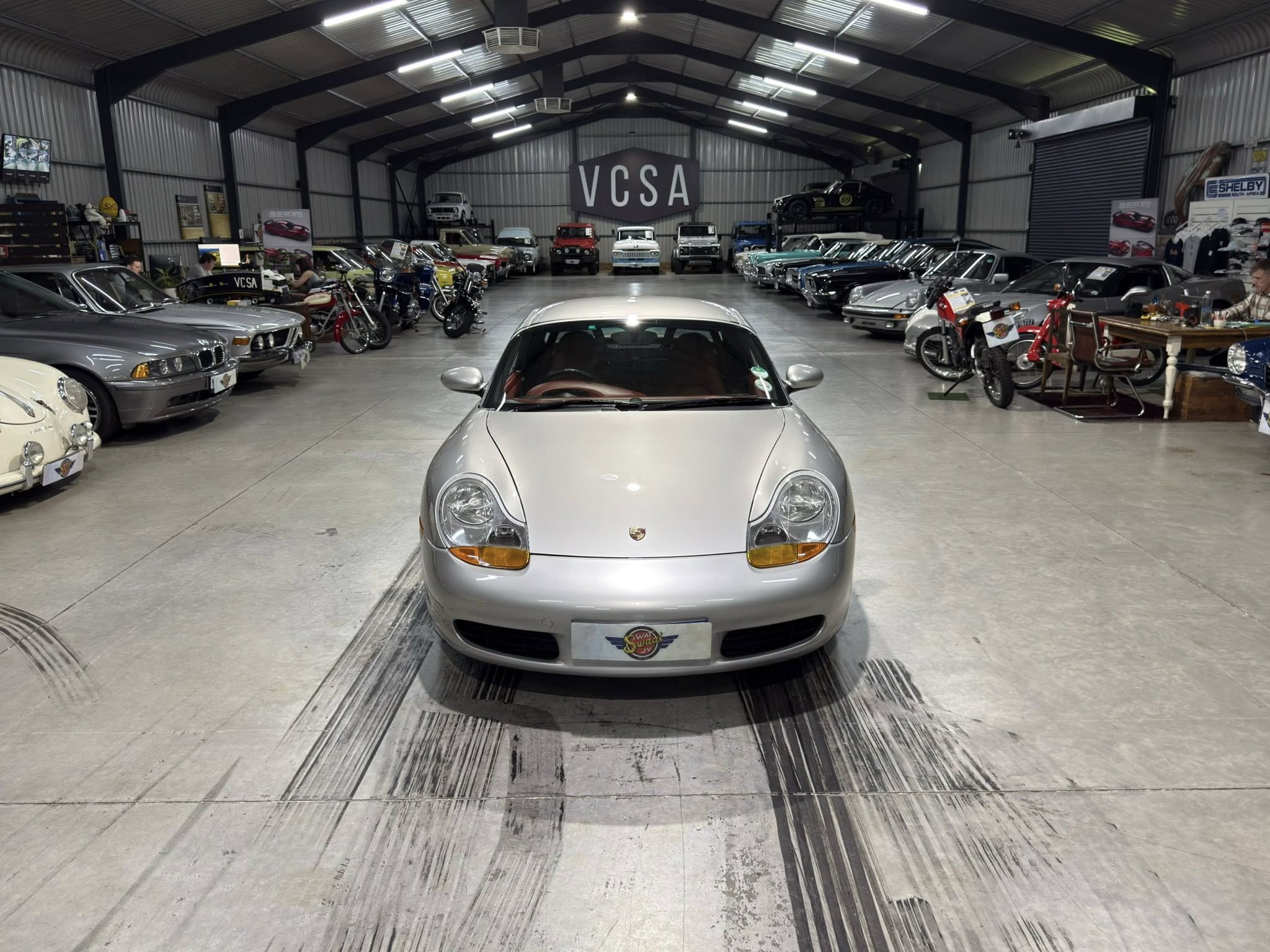 Front view of a silver Porsche in the center of a spacious showroom, flanked by rows of classic cars and motorcycles. A large VCSA sign is on the back wall.