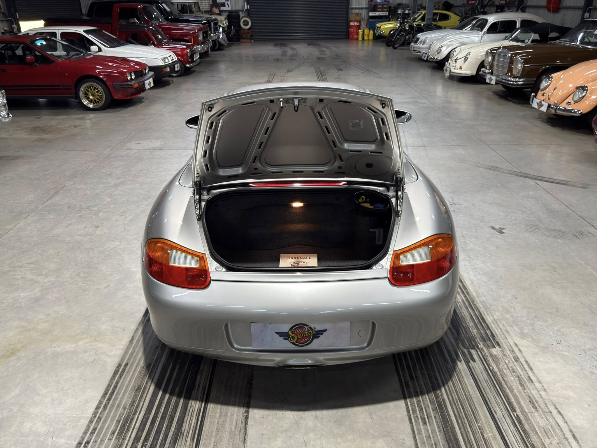 Silver sports car with its trunk open, seen from behind, in a showroom flanked by vintage cars on both sides.