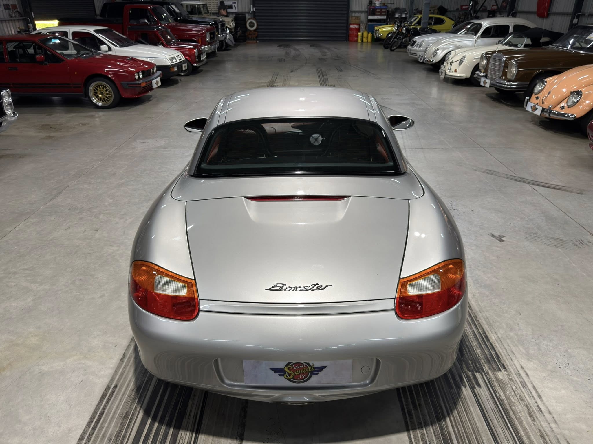 Rear view of a silver Porsche Boxster parked in a classic car showroom, with rows of vintage cars on both sides.
