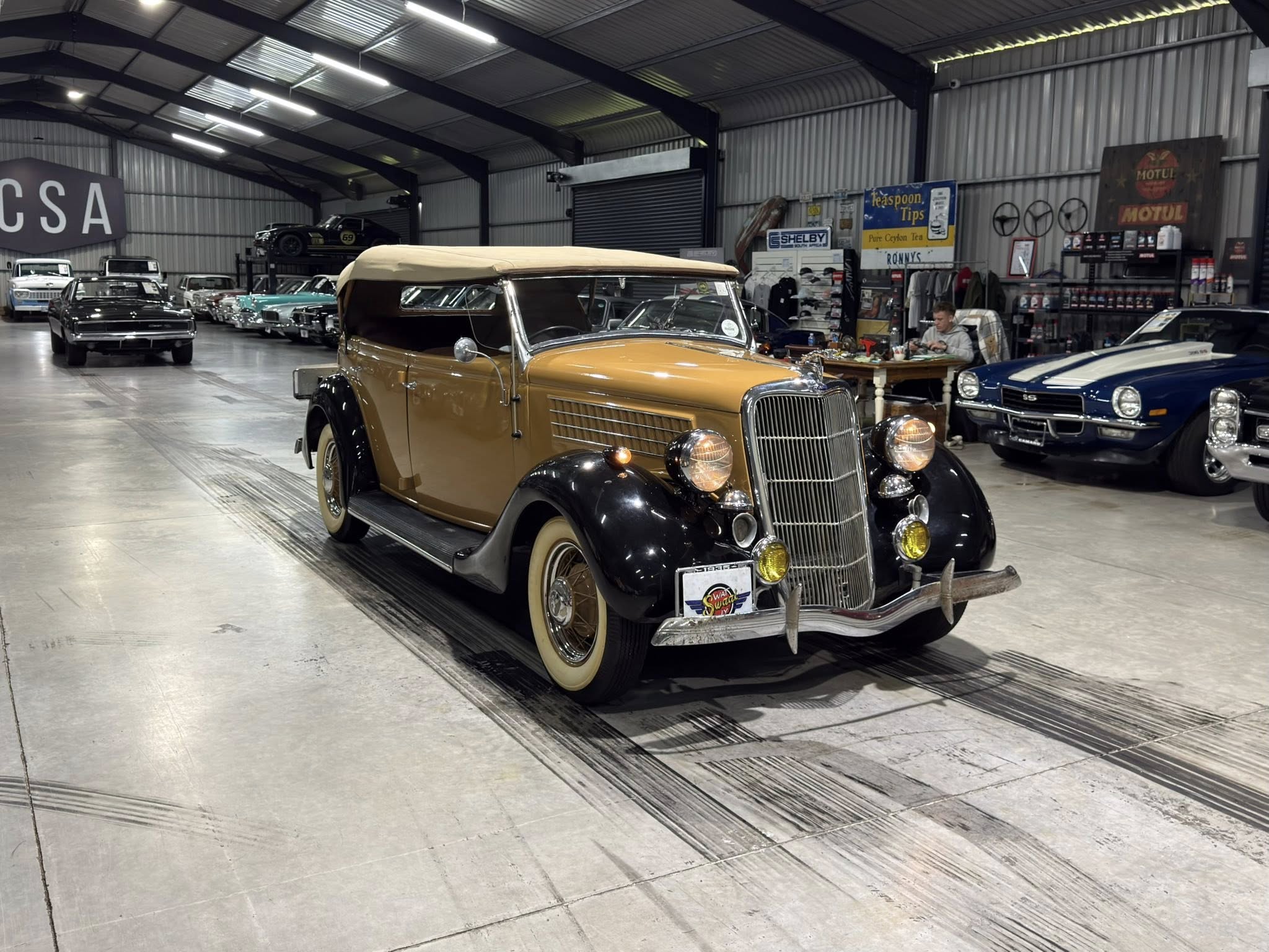 Indoor showroom with vintage cars; tan convertible in foreground and more classic cars visible in background.