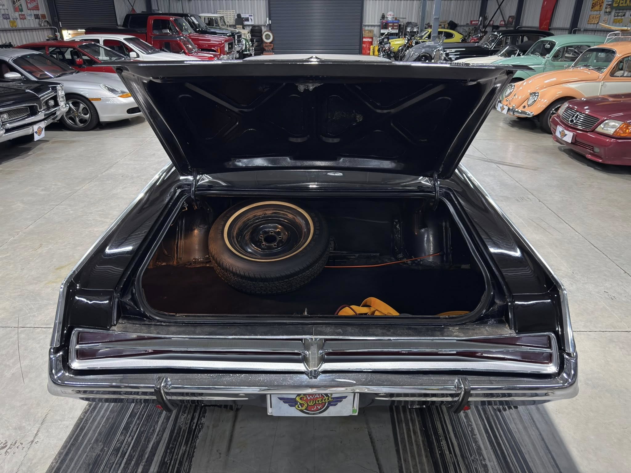 Open trunk of a glossy black vintage car in a showroom with many classic cars parked along the sides, spare tire visible inside the trunk.
