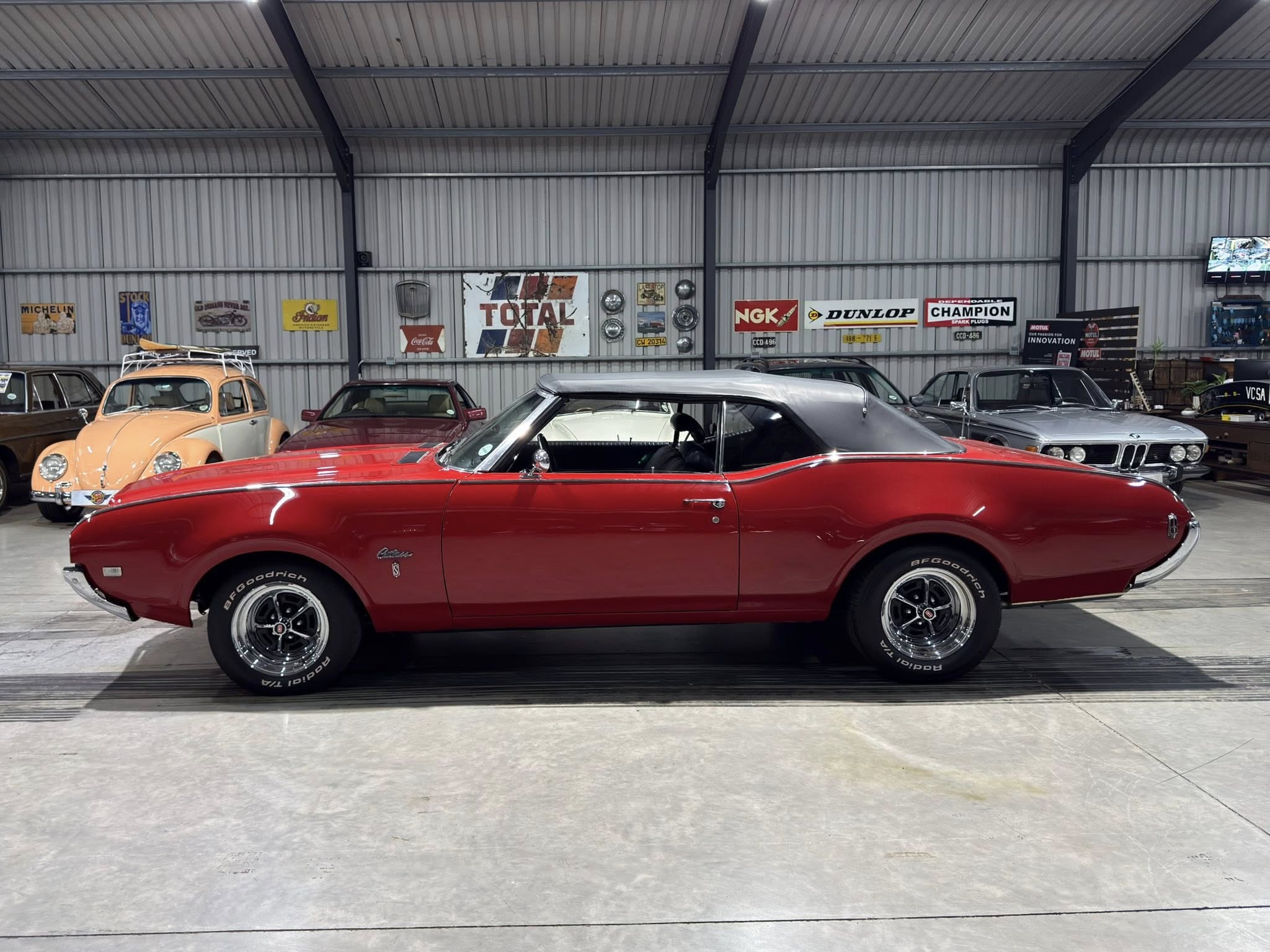 Red vintage muscle car in a showroom with other classic cars and retro signs on corrugated metal walls behind it.