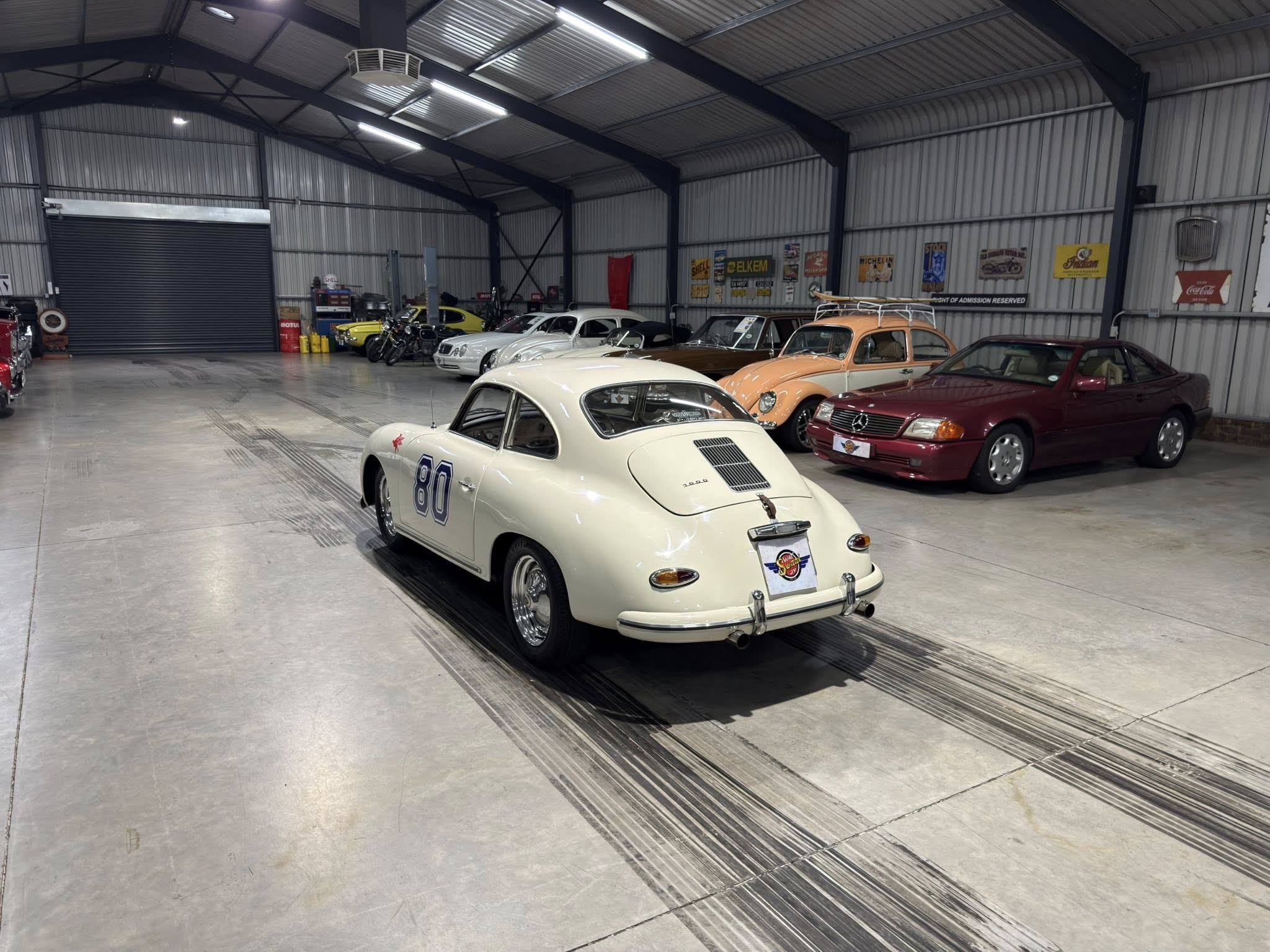 Interior of a spacious garage with vintage cars on display; a white racing car with number 80 in the foreground and classic cars lined along the wall in the background.