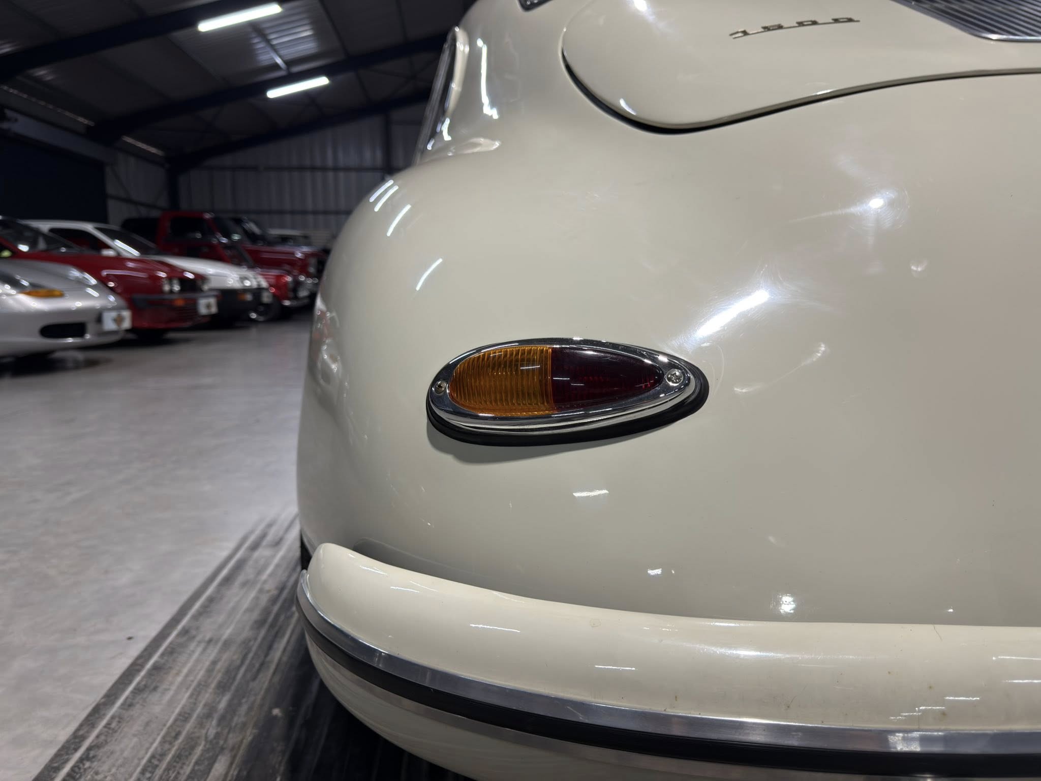 Close-up of a pale vintage car's front bumper with an amber signal light in a dim garage, with other cars in the background.