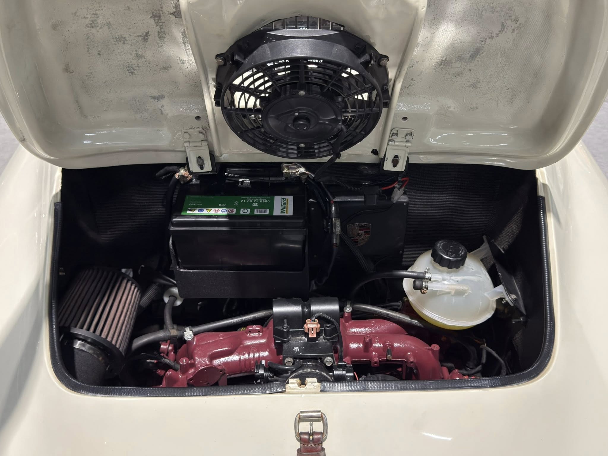 Under-hood view of a car engine bay: a circular cooling fan mounted at the back, with a green battery, hoses, and a coolant reservoir visible nearby.