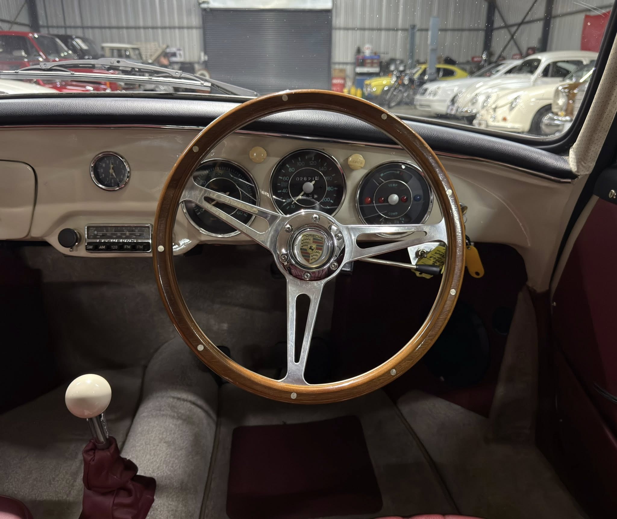 View from the driver’s seat of a vintage Porsche with a wooden steering wheel in a workshop/garage setting, other cars visible outside the windshield.