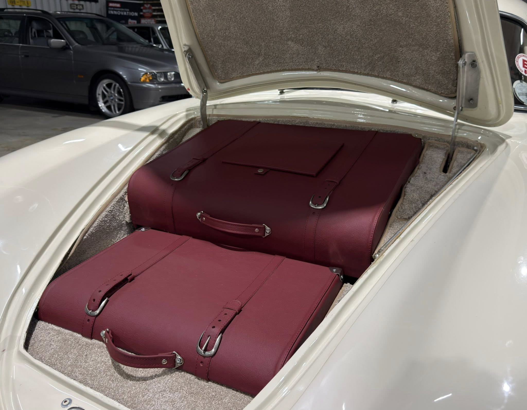 Open trunk of a white car revealing large red leather suitcases inside, beige carpeting, and other cars in a showroom background.