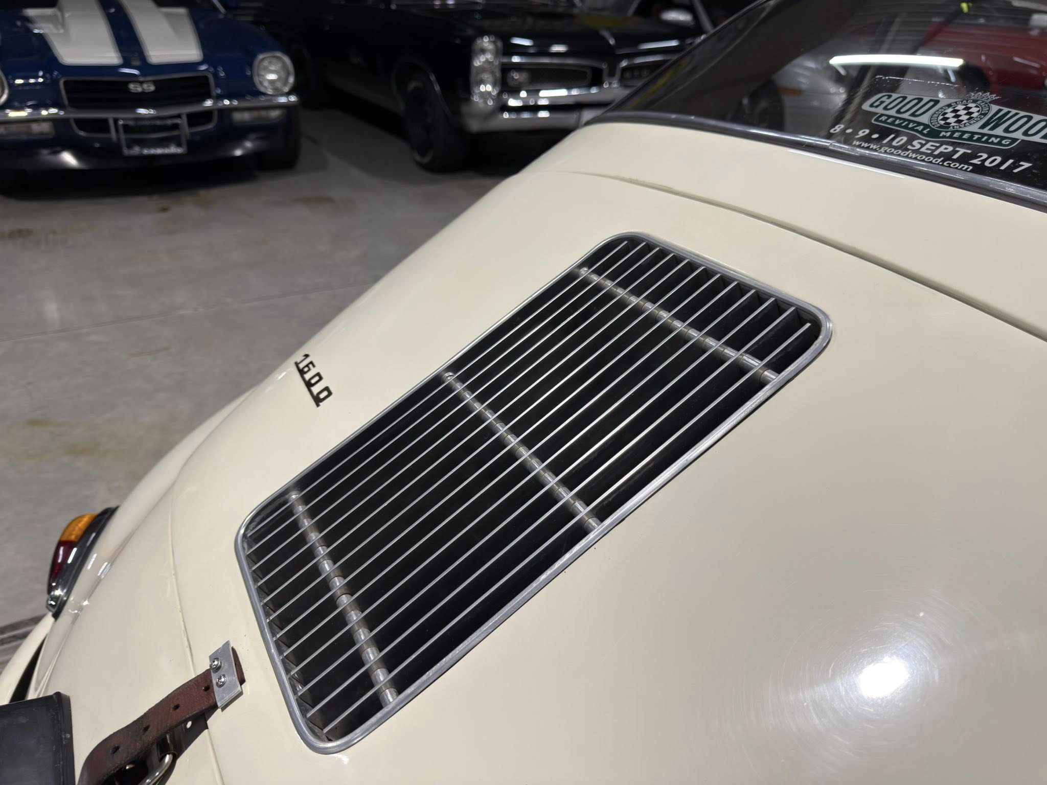 Beige vintage car hood with a chrome vent grille; background shows classic cars in a dim showroom.