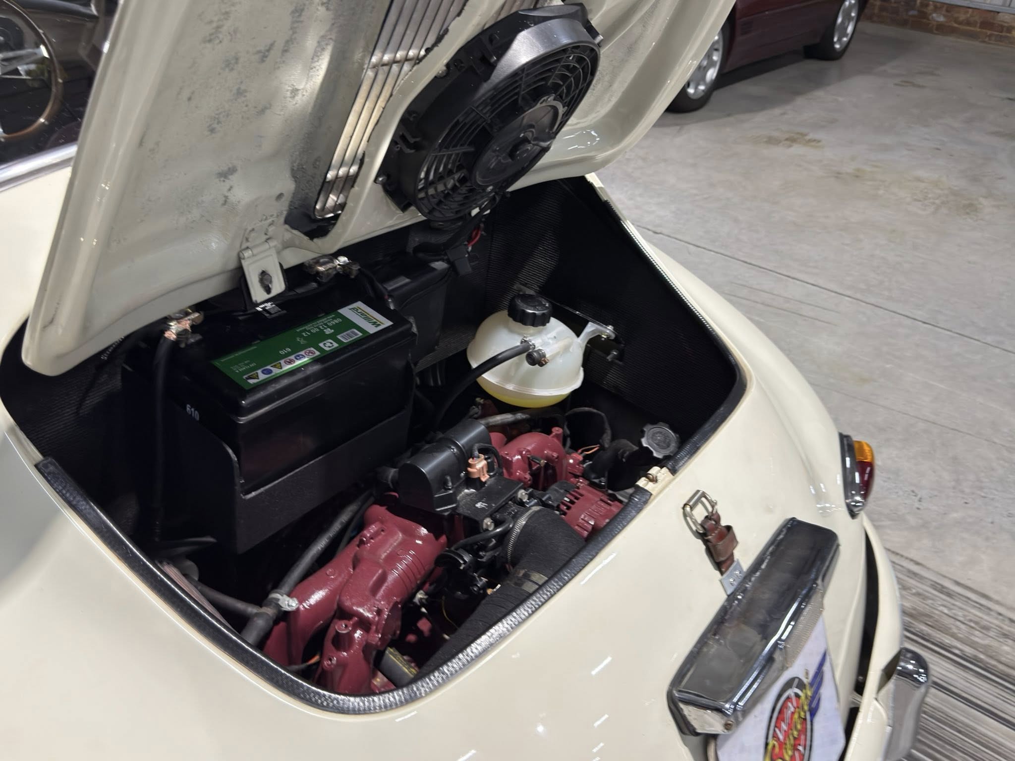 Open car rear hood shows engine bay with a battery, coolant reservoir, and cooling fan visible inside a light-colored trunk opening.