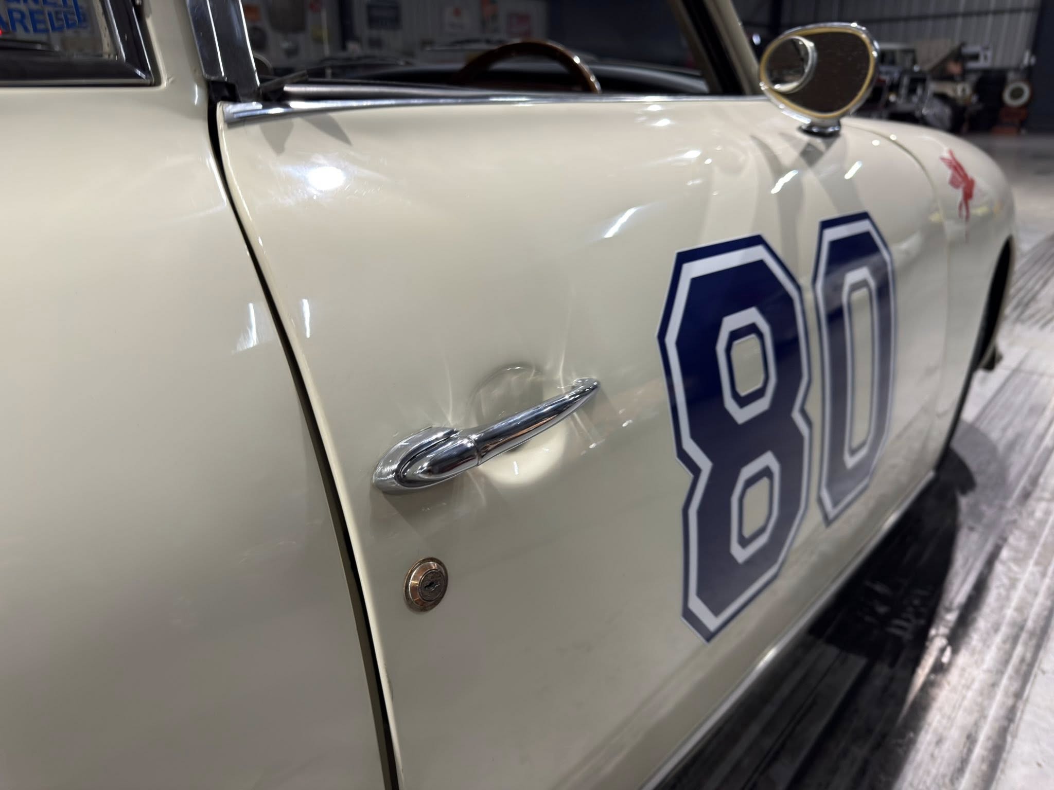 Close-up of a vintage cream-colored car door with chrome handle and blue number decals in a workshop setting.