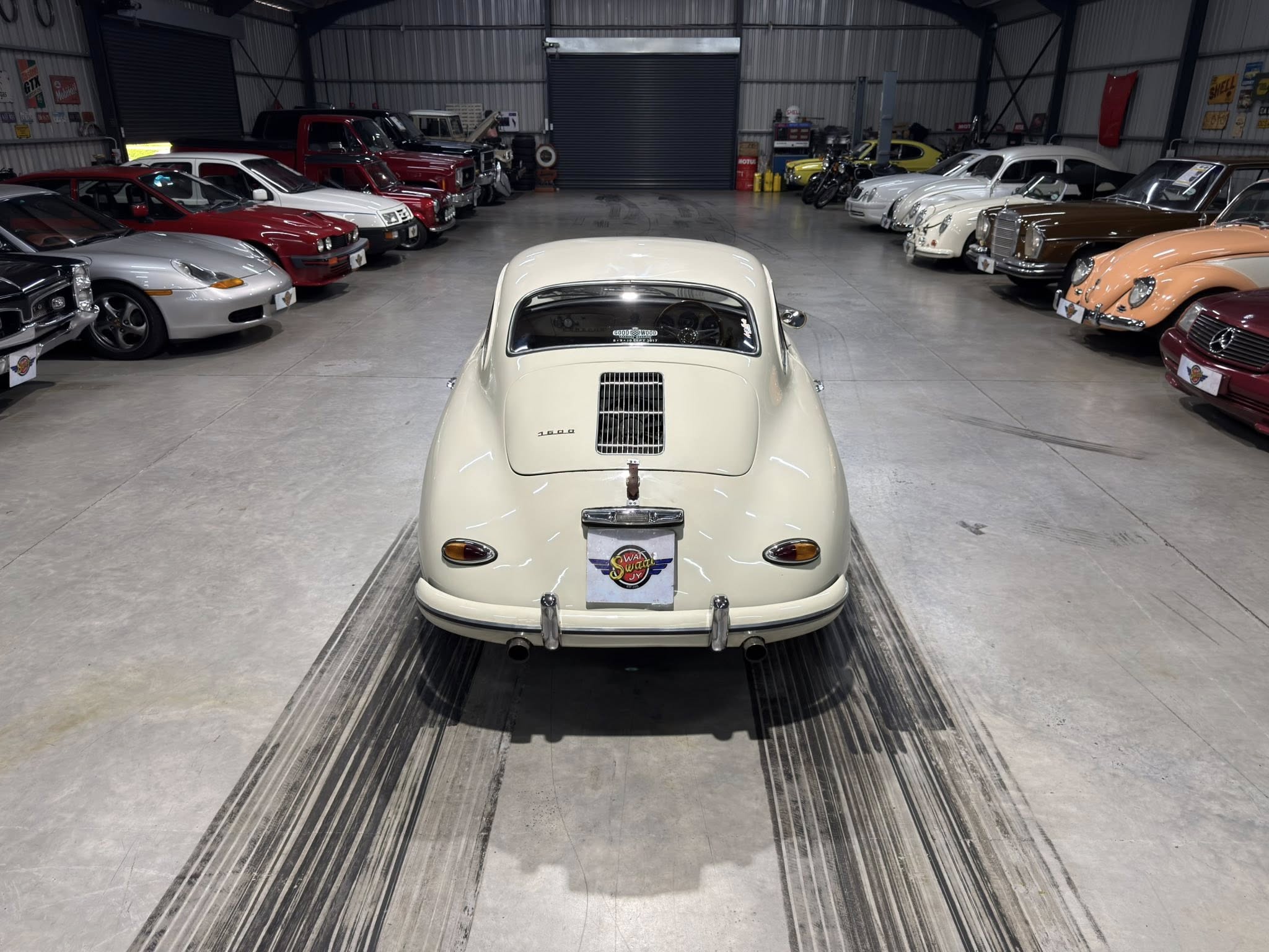 Inside a spacious showroom filled with classic cars, viewed from behind a cream-colored vintage car at the center.