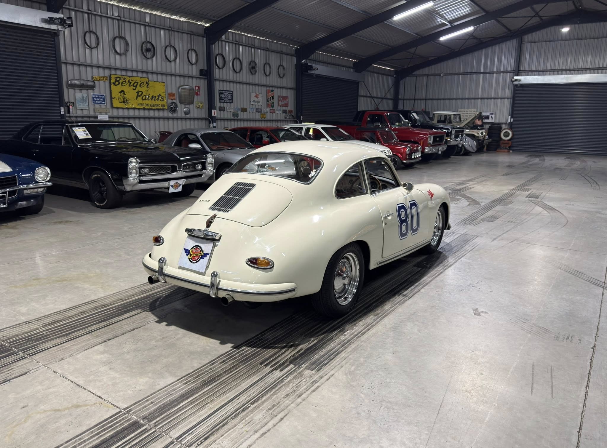 Cream Porsche 356 with the number 80 in the foreground inside a metal-walled garage filled with other vintage cars to the left and background.