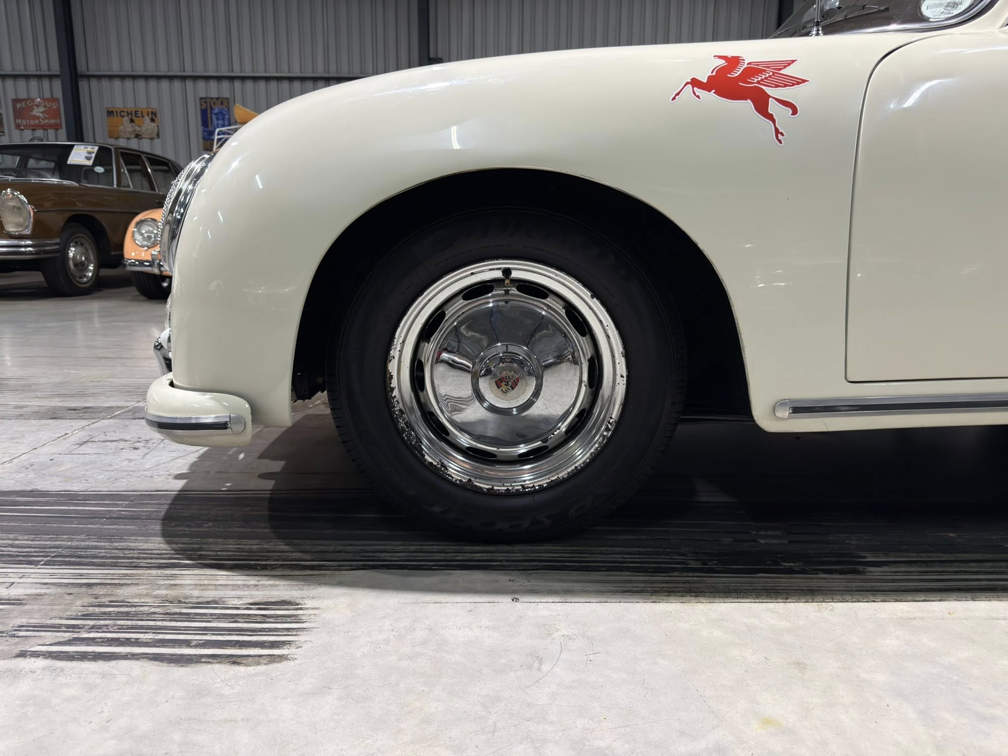 Close-up of a cream-colored vintage car's front wheel and fender with a red Pegasus emblem on the hood, in a showroom setting