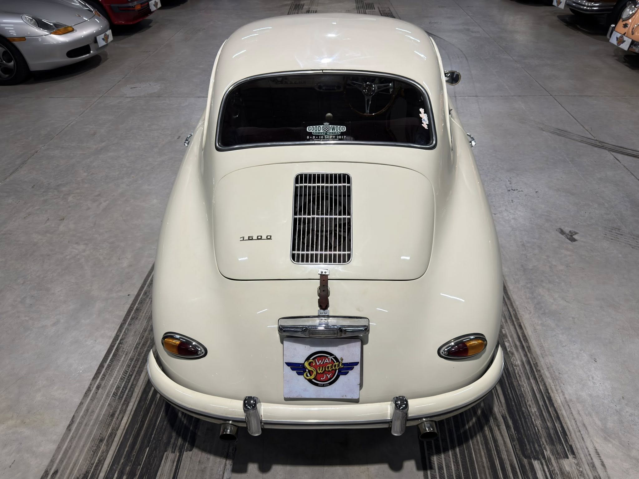 Rear view of a cream-colored vintage car in a showroom, featuring a chrome bumper, small tail lights, and a prominent grille/vent on the hood edge with 1500 badge nearby.
