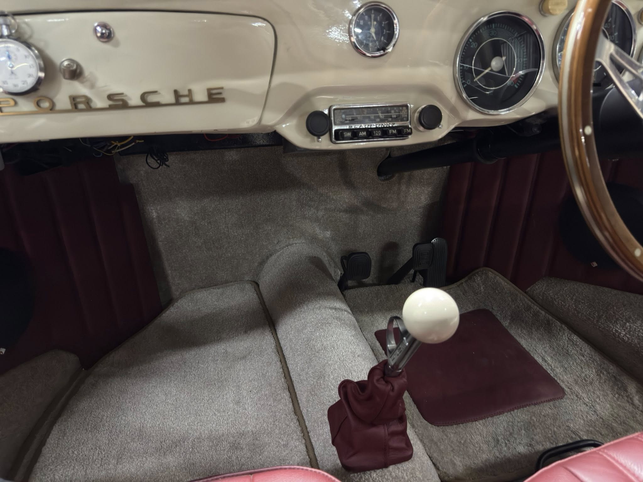 Vintage Porsche dashboard interior with beige color, showing analog gauges, center radio, and wooden steering wheel; shifter with white knob and beige carpeted floor mats visible.