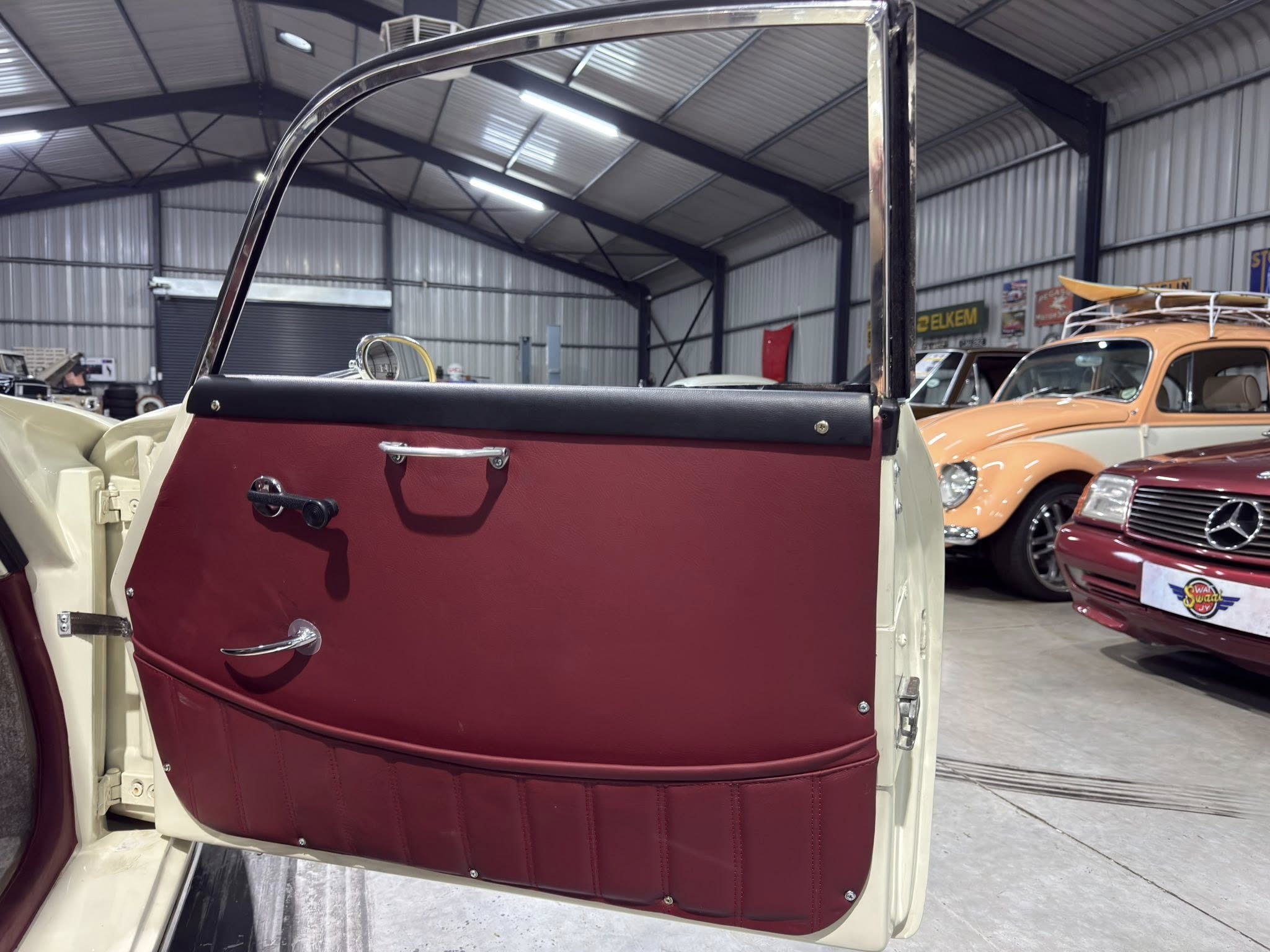 Open passenger door of a cream-colored vintage car with burgundy interior, chrome trim visible inside a showroom with other classic cars in the background.