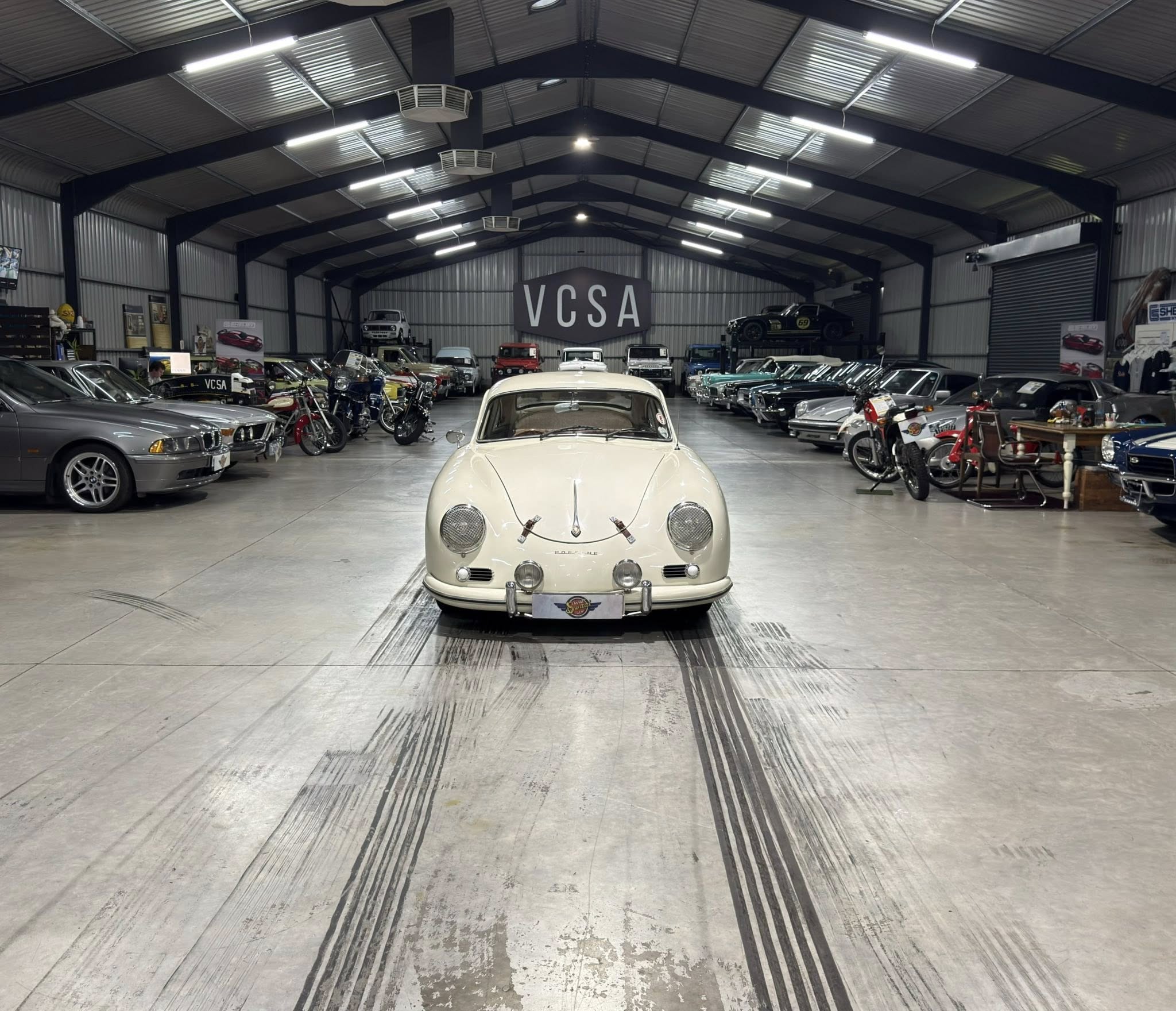 Indoor car showroom filled with classic cars; cream vintage car centered in the foreground, rows of vehicles lining both sides, and a large VCSA banner in the back.