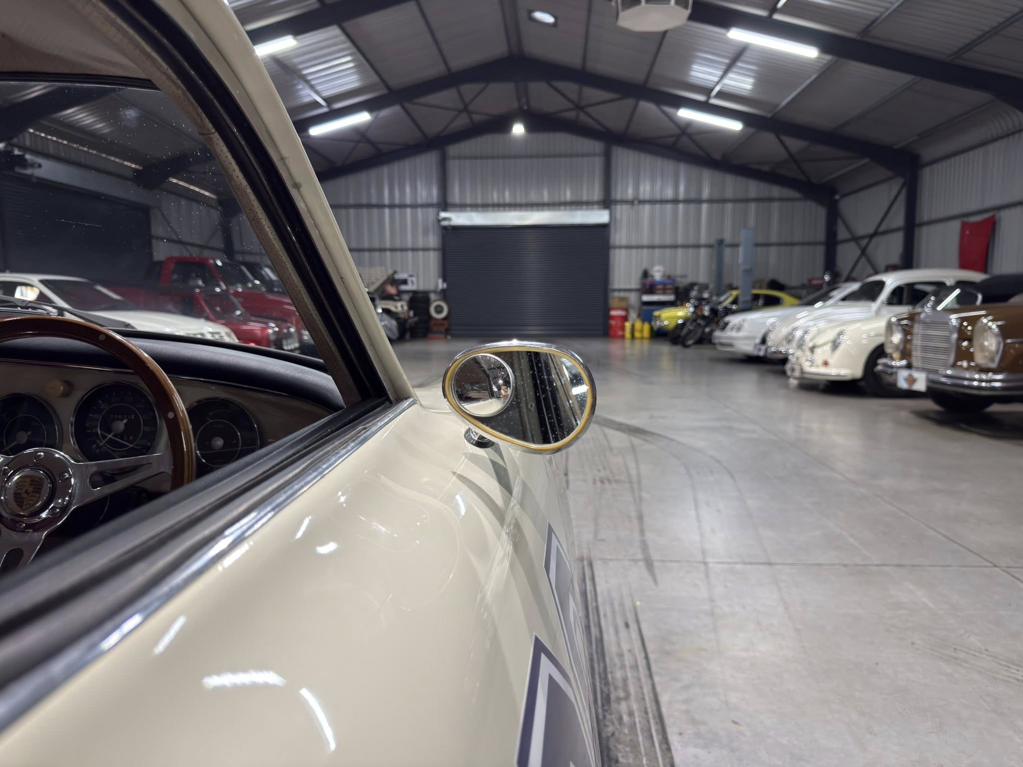 Front-seat view of a classic car interior, showing the steering wheel and dashboard, looking into a spacious showroom with vintage cars lined up along the right.