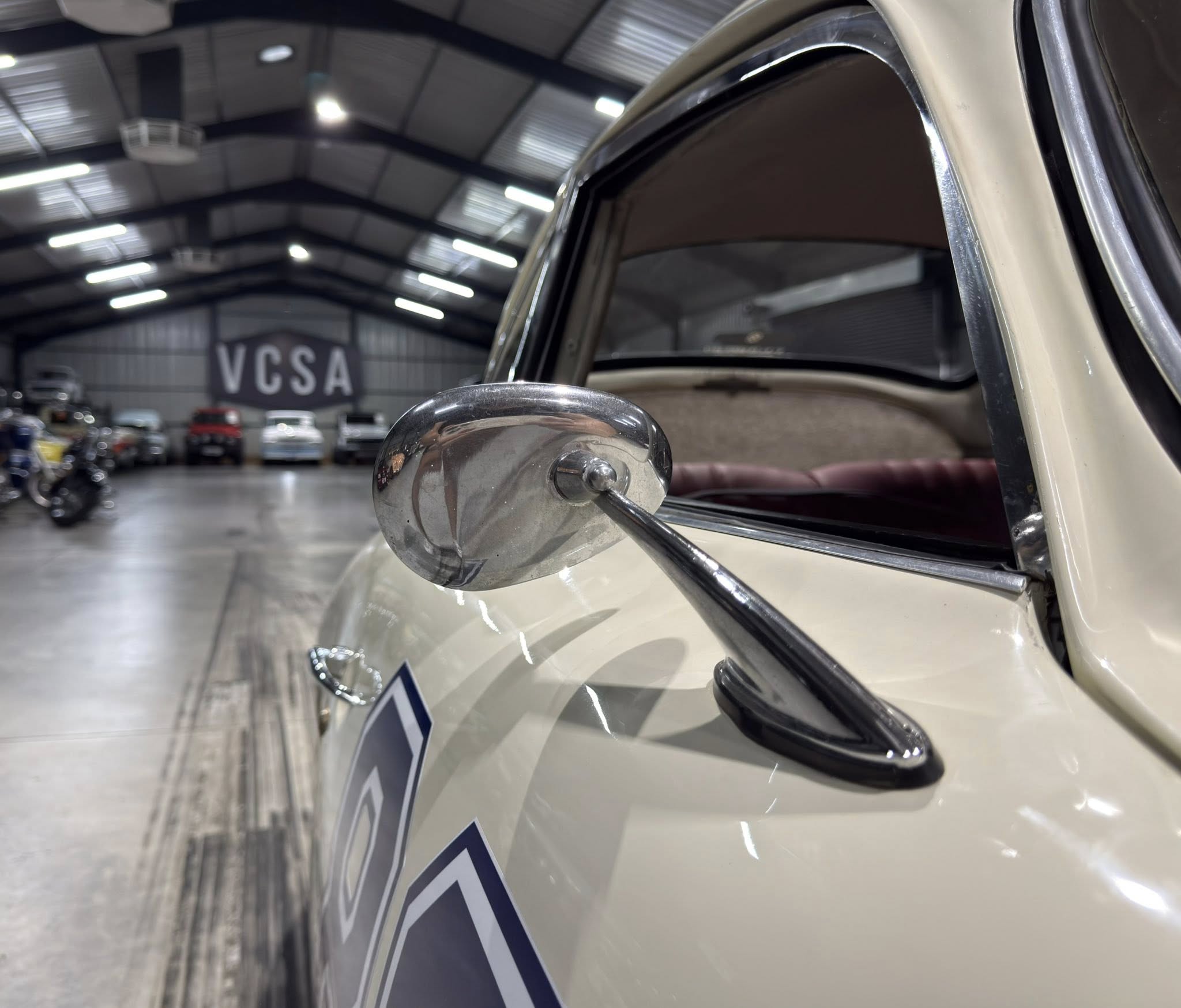Close-up of a chrome side mirror on a cream vintage car, indoor showroom with other cars in the background.