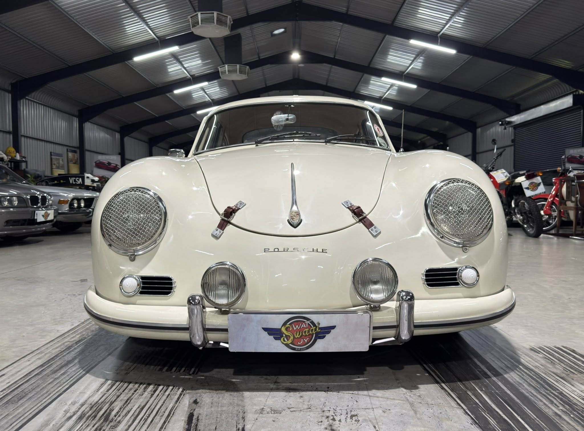 Front view of a vintage white Porsche in a showroom, with other cars and motorcycles in the background, indoors under metal beams.