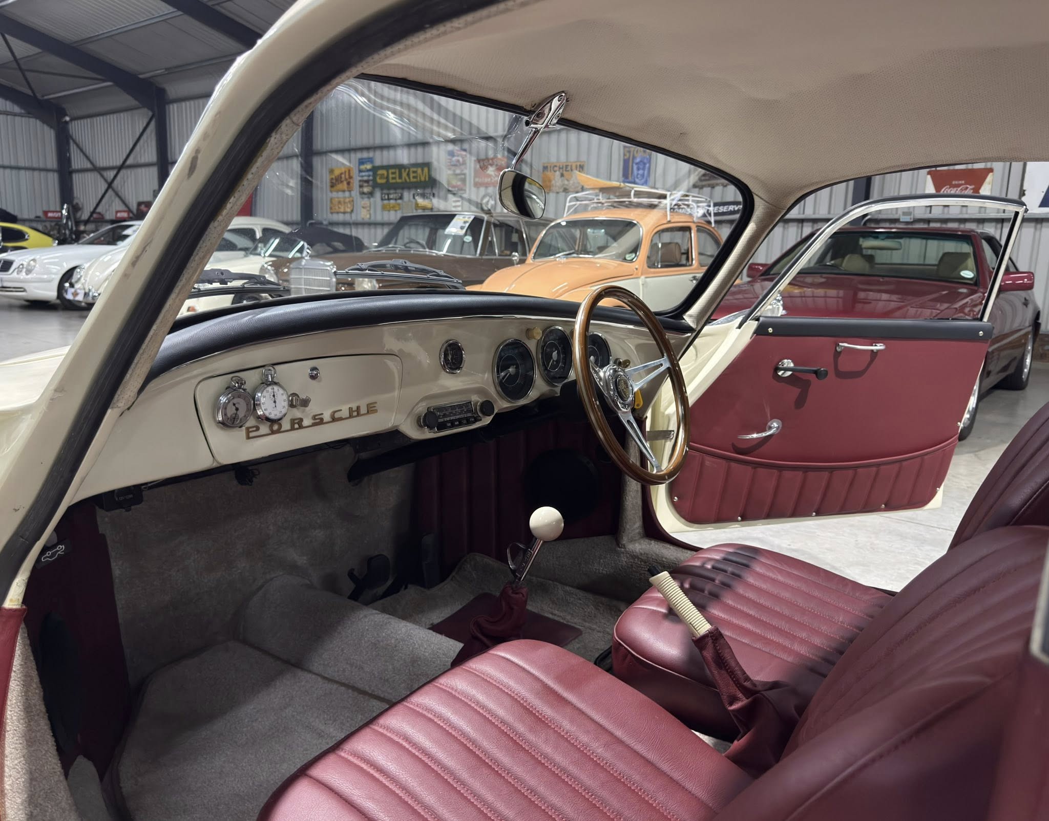 Interior view of a classic Porsche with burgundy leather seats, cream dashboard, and a wooden steering wheel in a showroom of vintage cars.