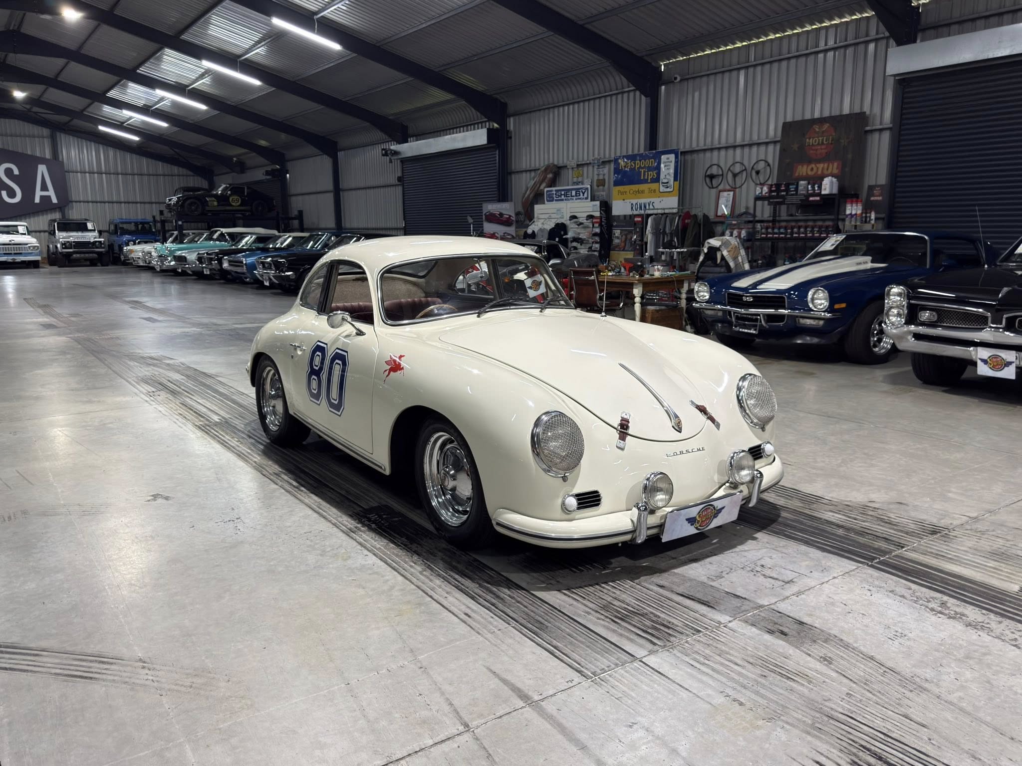 White vintage Porsche 356 coupe in a spacious showroom with other classic cars lined up along the wall, indoor lighting overhead.