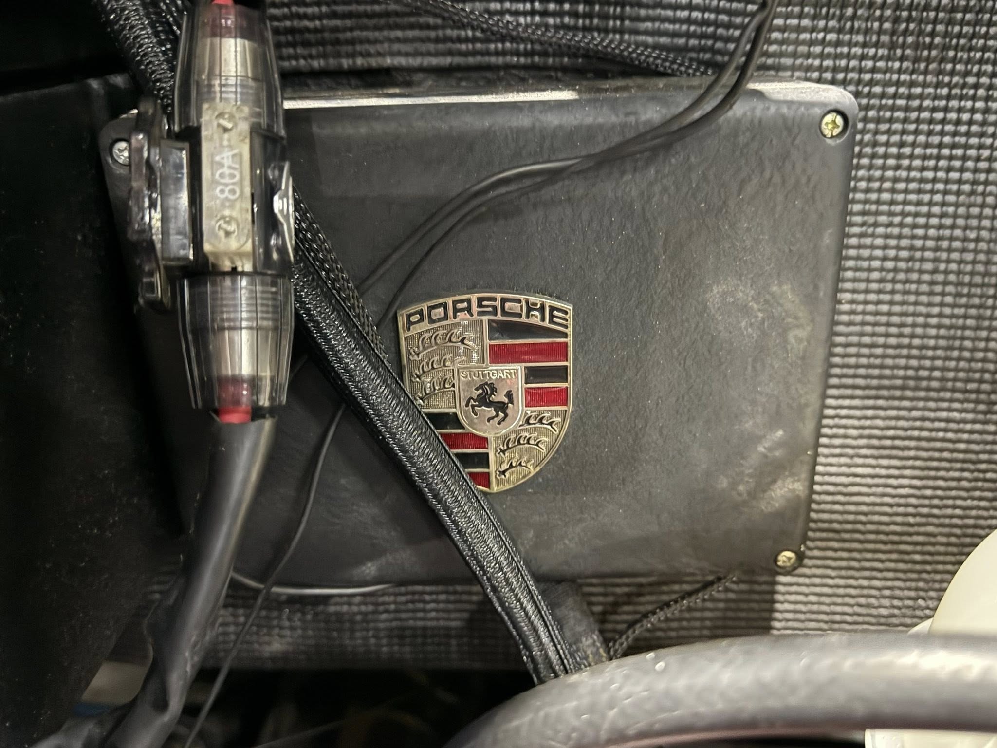 Close-up of a Porsche crest badge on a black car interior panel, with a textured surface and steering wheel rim in the foreground.