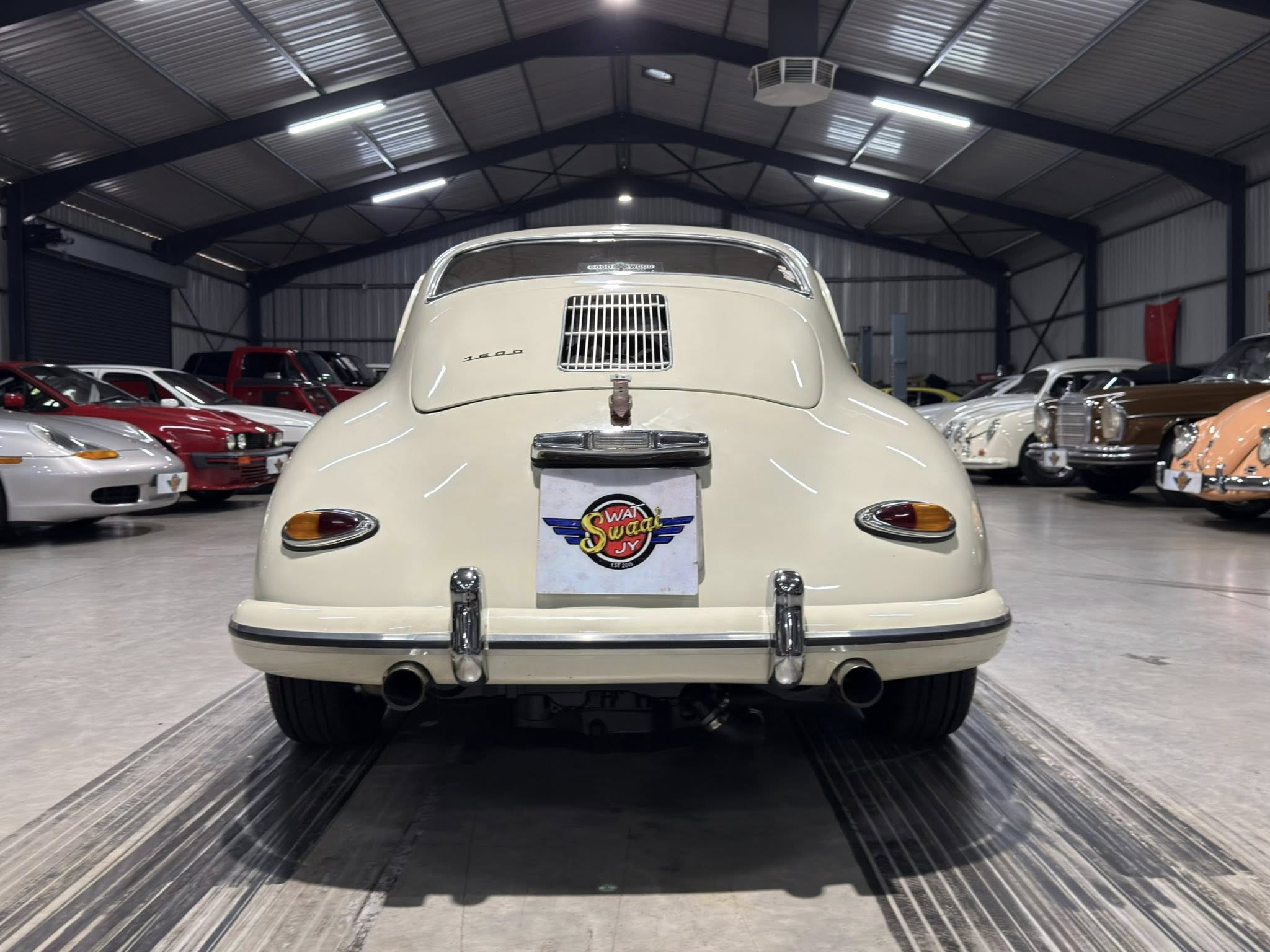 Rear view of a cream-colored classic car in an indoor showroom, with other vintage cars parked along the sides.