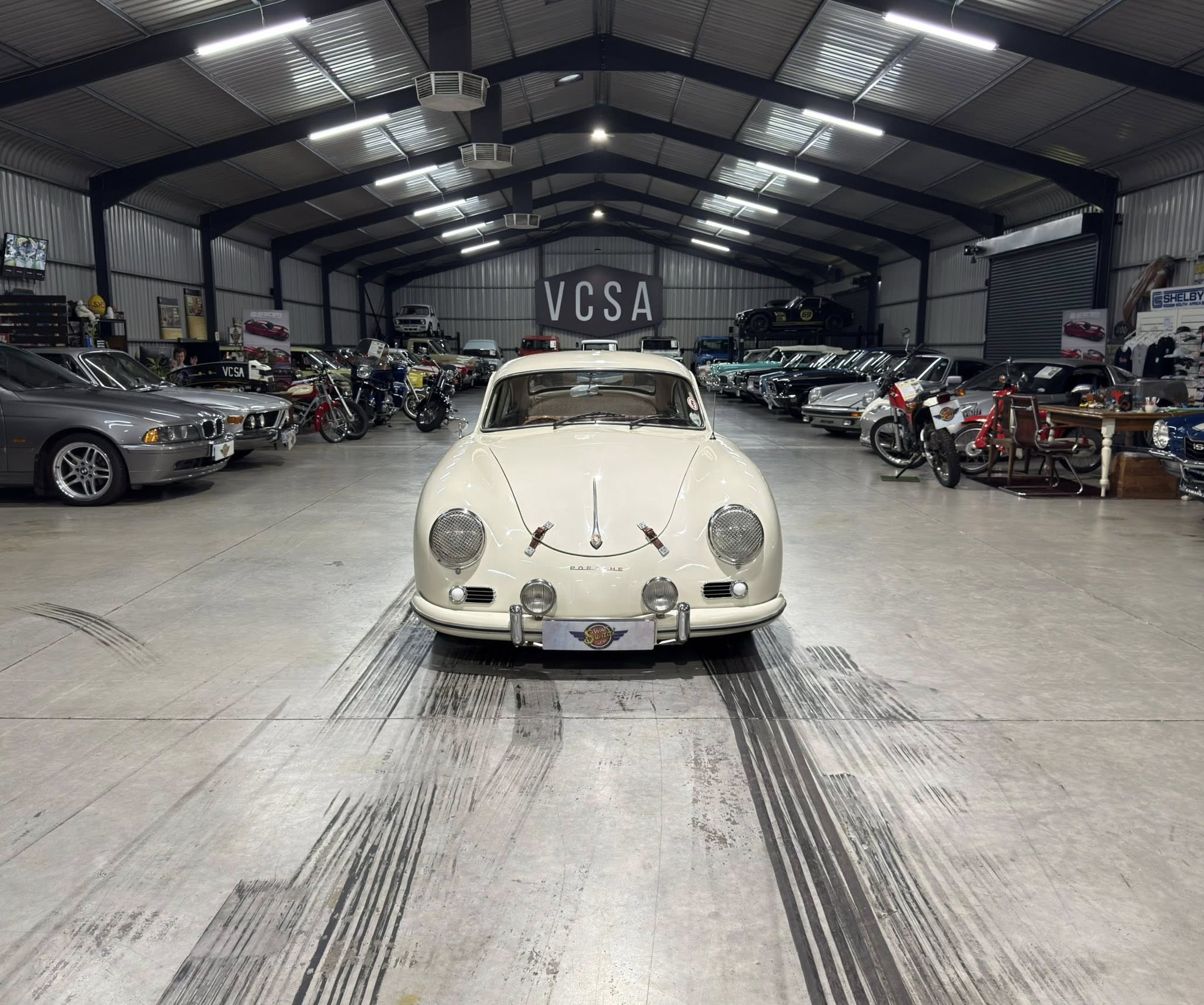 Indoor car showroom filled with vintage cars and motorcycles; white classic car centered in the foreground with a VCSA banner in the back hall.