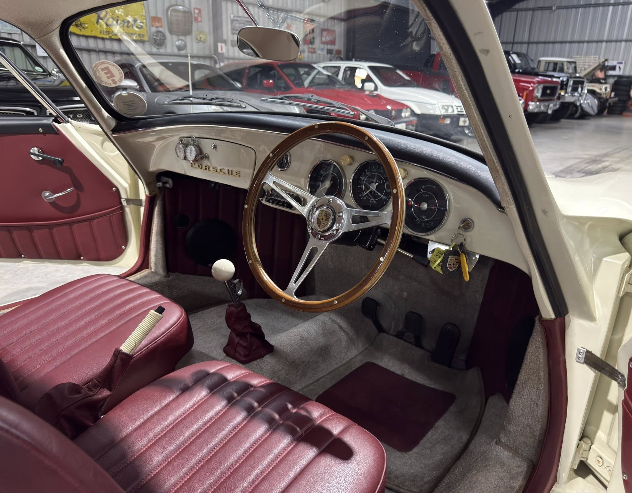 Inside a vintage Porsche cabin with burgundy seats, wood steering wheel, and classic gauge cluster; other classics visible through windshield.