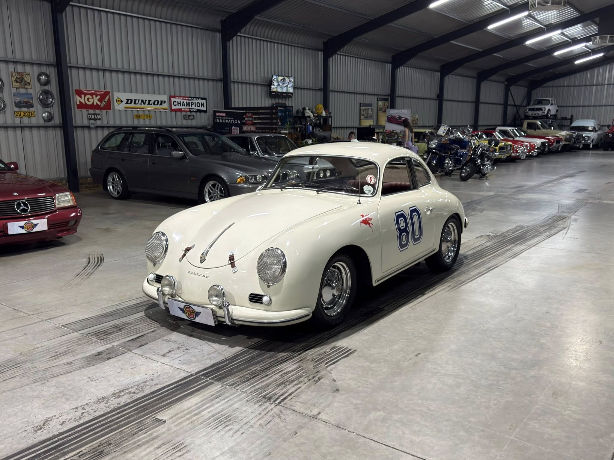 Indoor showroom of classic cars: prominent white vintage Porsche 356 with race number 80, surrounded by other antique cars and motorcycles.
