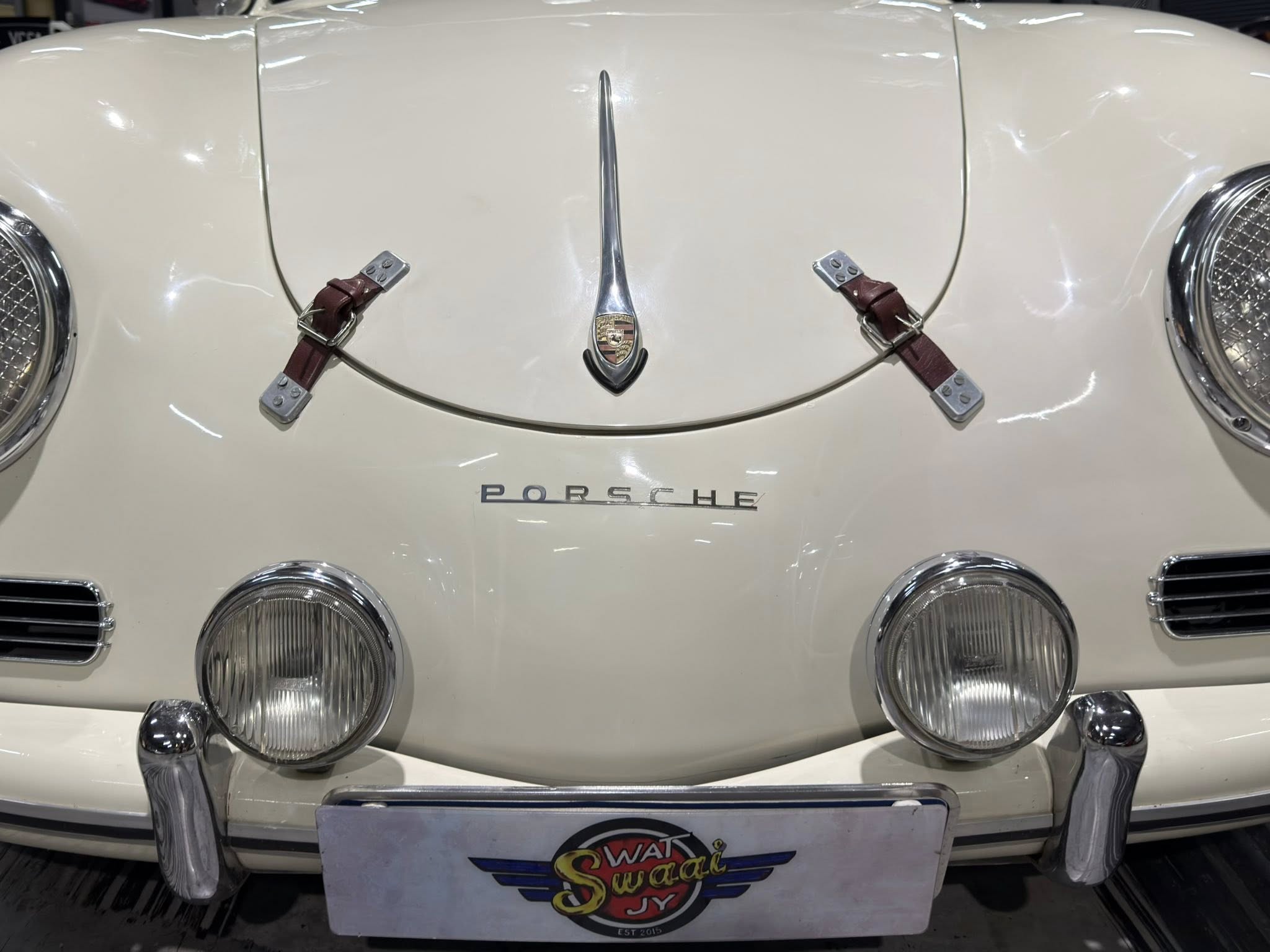 Front close-up of a white vintage Porsche with leather hood straps and central crest emblem between the straps and round headlights visible on either side.
