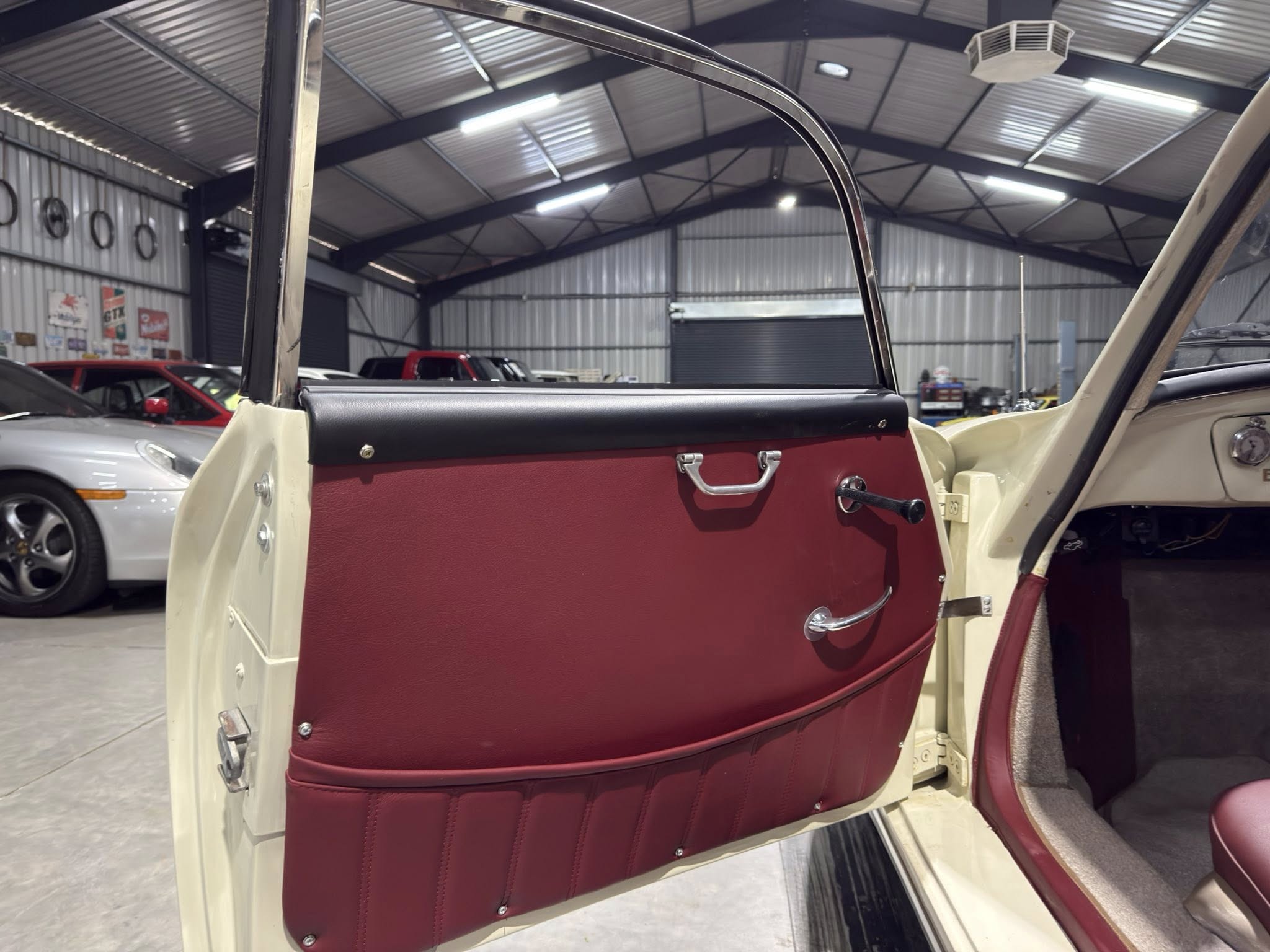 Open car door exposing the red leather interior of a vintage vehicle, inside a large workshop/garage with other cars in the background.