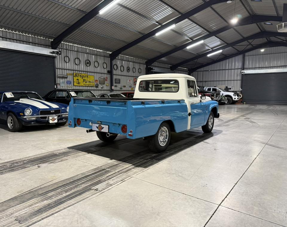 Blue vintage pickup truck with a white cab on display inside a warehouse-style showroom among other classic cars.