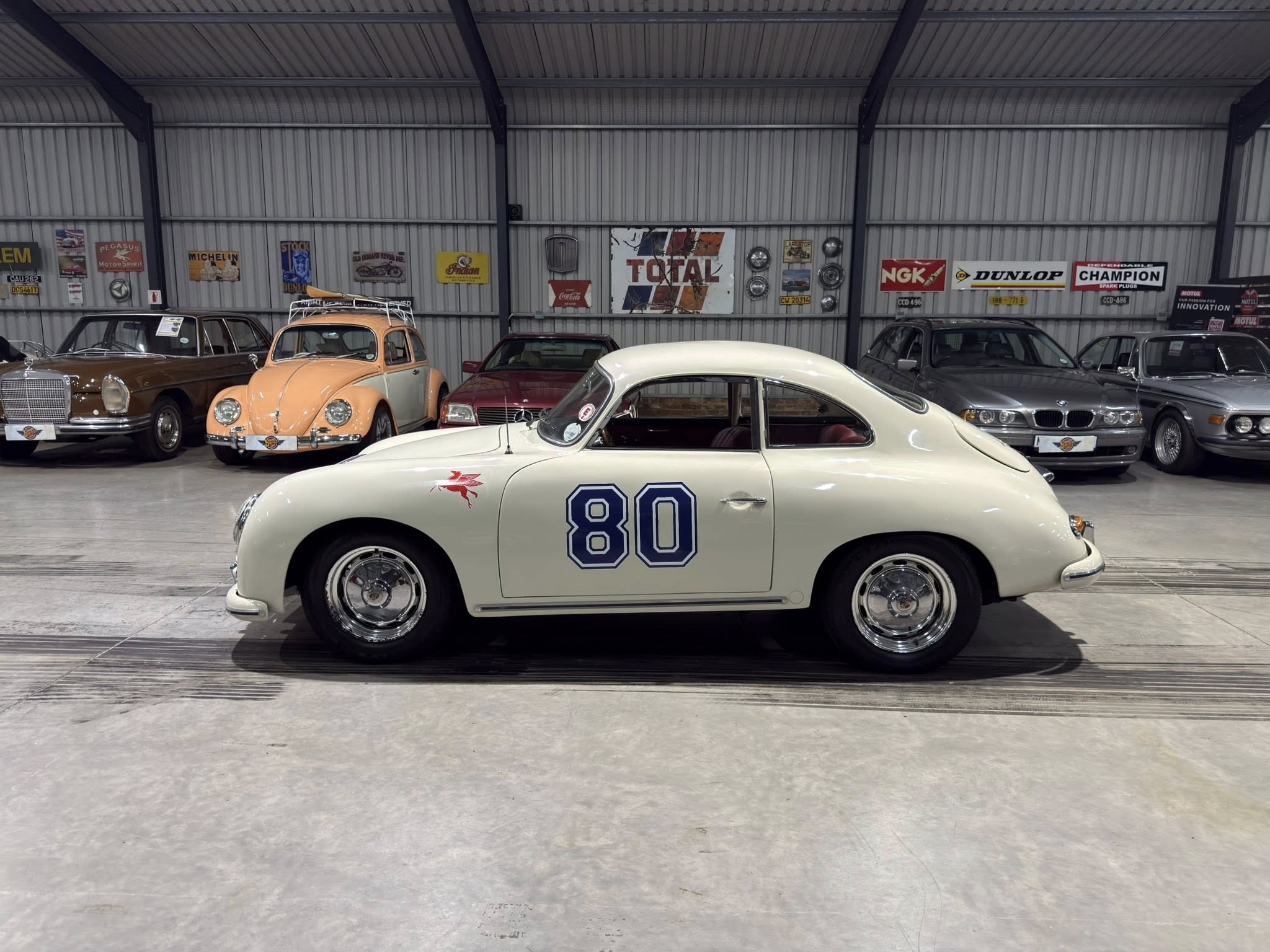 White vintage racing coupe in the foreground with the number 80, surrounded by other classic cars in a large showroom/garage with retro signs on the wall.