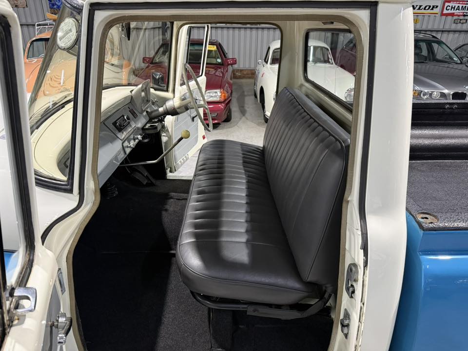 Interior of a vintage pickup: black vinyl bench seat, steering wheel, and cream-colored dashboard visible through open door. White exterior, blue truck bed in view.