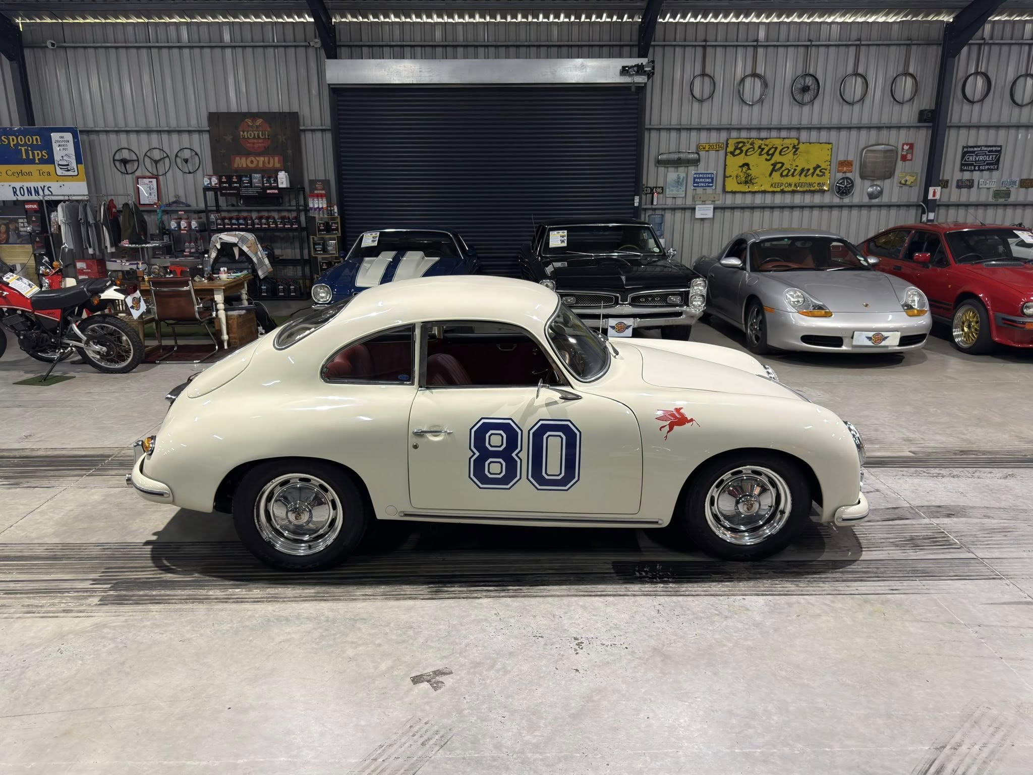 White vintage race car with number 80 parked indoors among other classic cars in a showroom/garage setting.