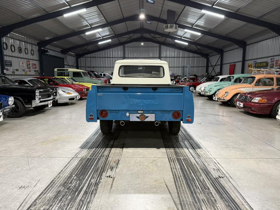 Blue vintage pickup seen from the rear inside a large car showroom filled with classic cars on both sides.