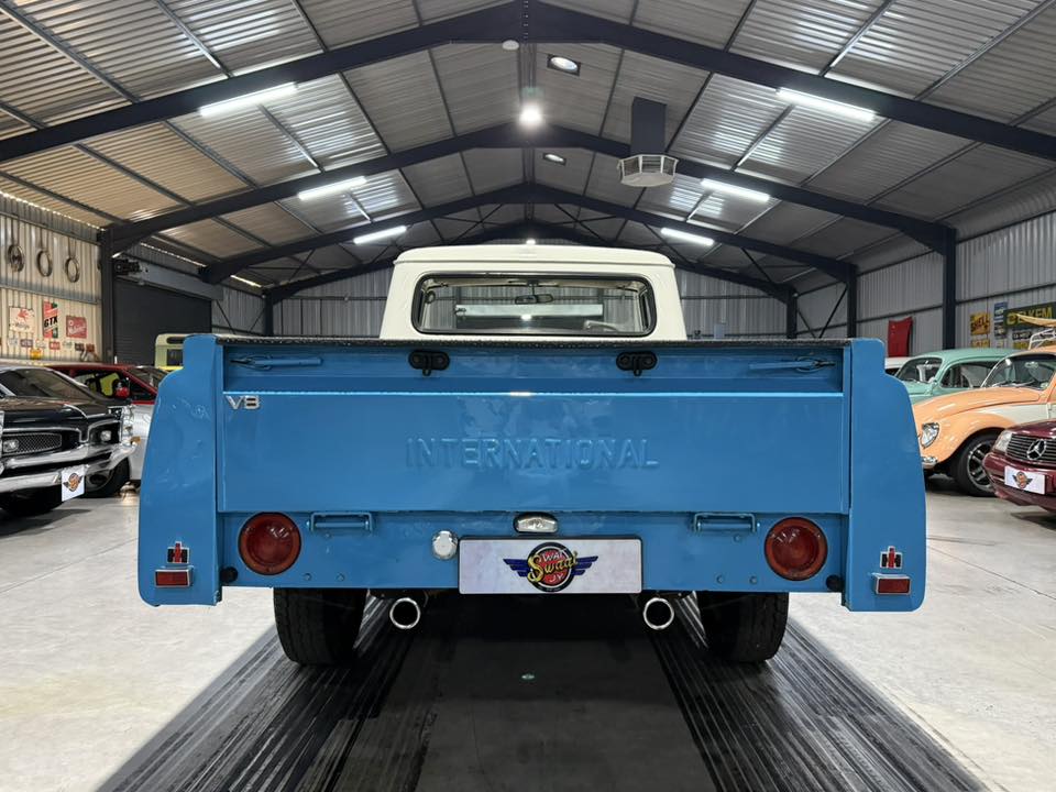 Rear view of a bright blue vintage International pickup truck inside a showroom, white cab roof visible, twin exhausts and red circular taillights.