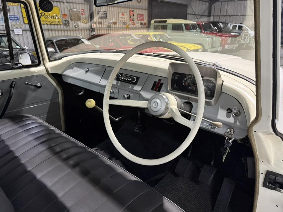 Interior of a vintage car: cream dashboard, large white steering wheel, manual gear shifter, and black bench seat in a showroom setting with other classic cars outside the windshield