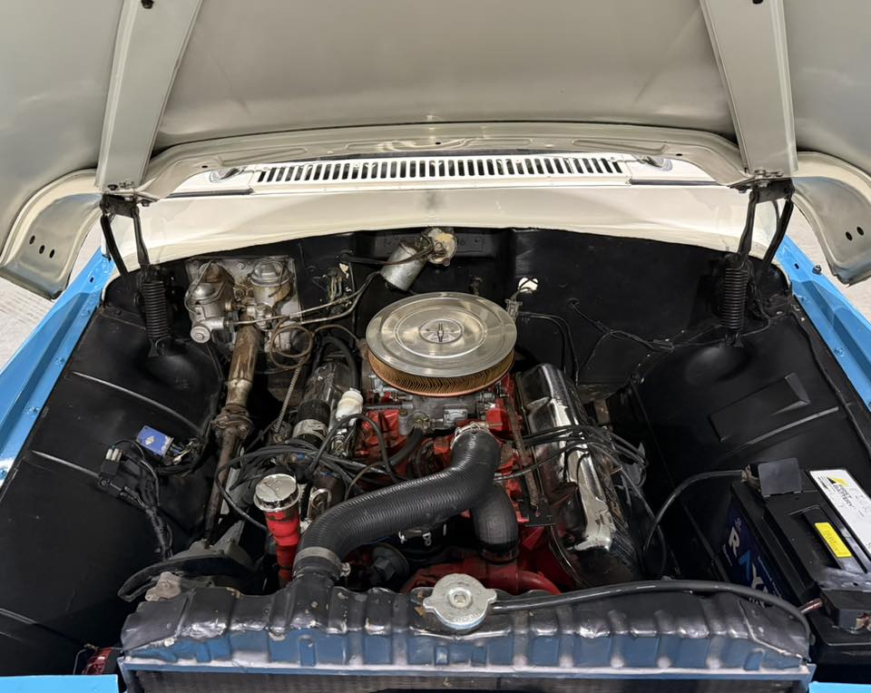 Open hood view of a classic car engine bay featuring a circular chrome air filter atop a red engine block and blue body panels in the foreground without trim.