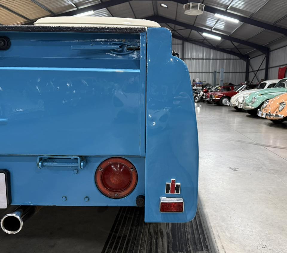 Blue vintage car rear end in a showroom with other classic cars in the background, indoors under bright lighting.