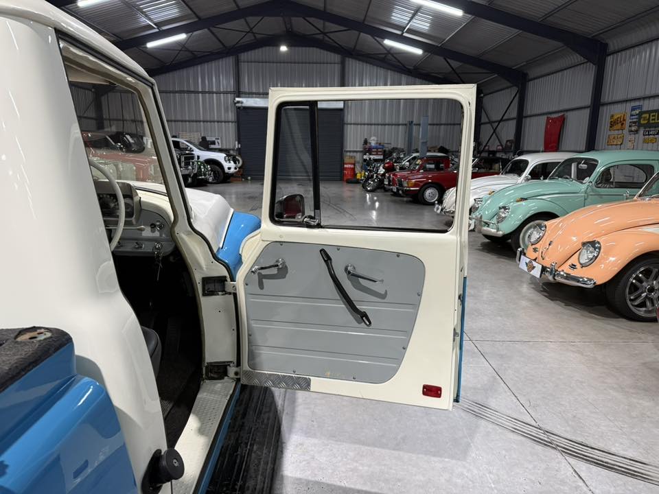 Open passenger door of a vintage white-and-blue car, showing the gray interior door panel and step plate, in a classic car showroom with other colorful retro cars in the background.