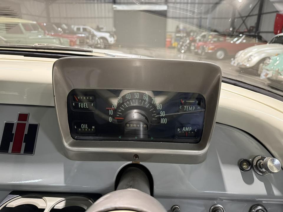 Dashboard of a vintage car with a central speedometer and gauges for fuel, oil, temperature, and amperage; showroom with other cars visible outside the windshield.