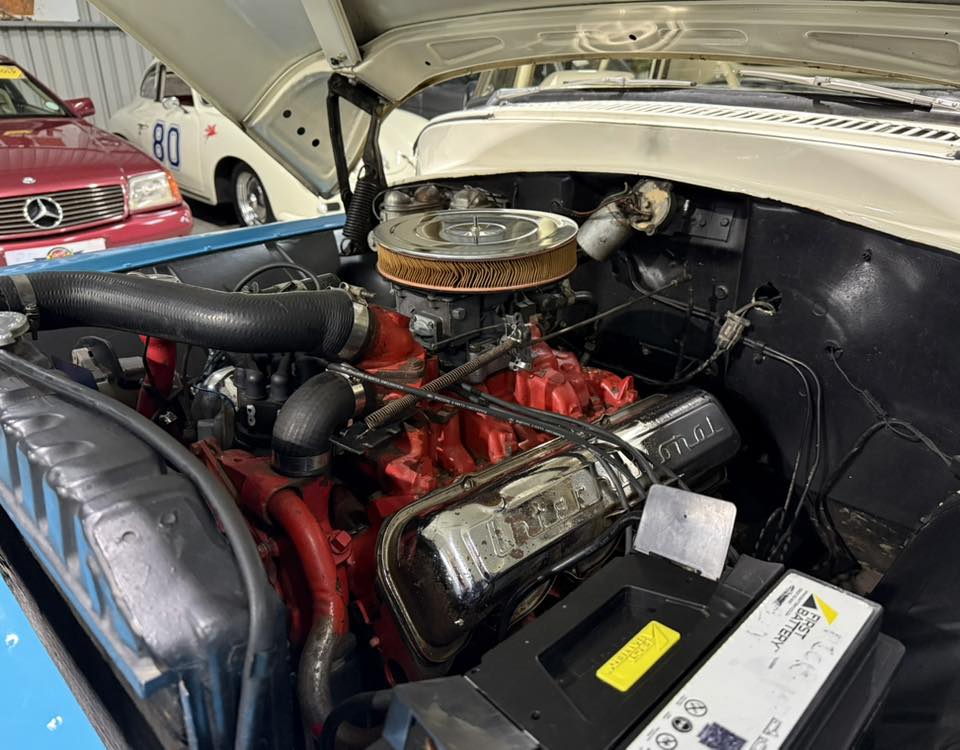 Open-hood view of a classic car engine: red engine block, chrome valve covers, round air filter, battery in the foreground, with race cars visible in the background.