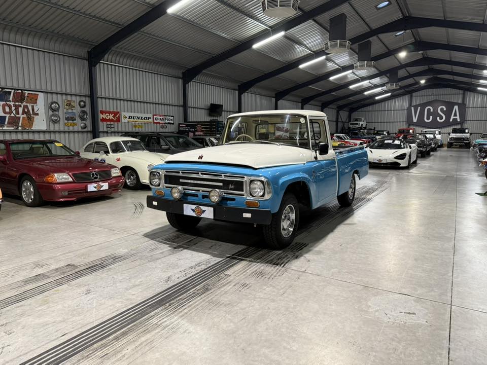 Blue and white vintage pickup parked in a spacious indoor showroom with classic cars on both sides.