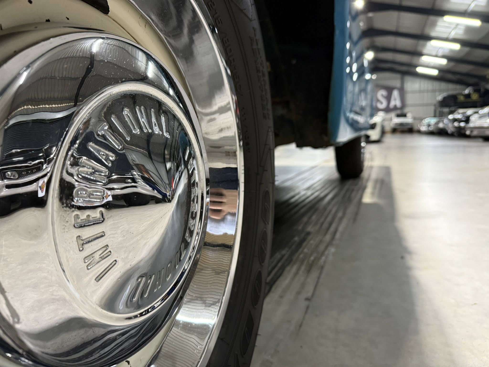 Close-up of a chrome car wheel and tire in a showroom with other cars blurred in the background.